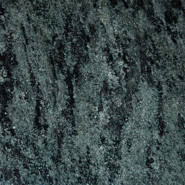 A close-up image of a dark green and black granite texture, showcasing its natural, mottled pattern and rough surface. Irregular streaks of black contrast with the deep green, accented by tiny lighter specks, emphasizing the stone's organic beauty suitable for memorials.